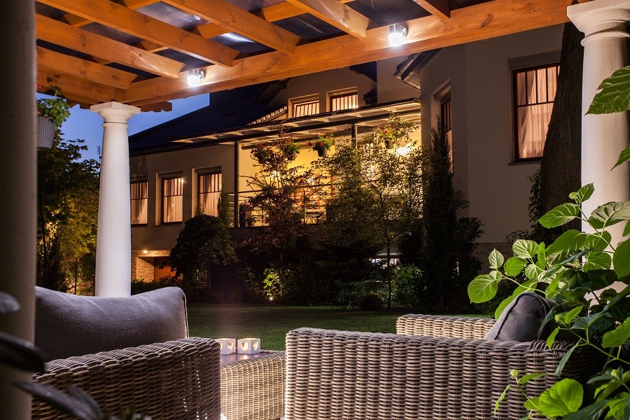 Sophisticated wicker chairs under a pergola with home in background illuminated by landscape lighting